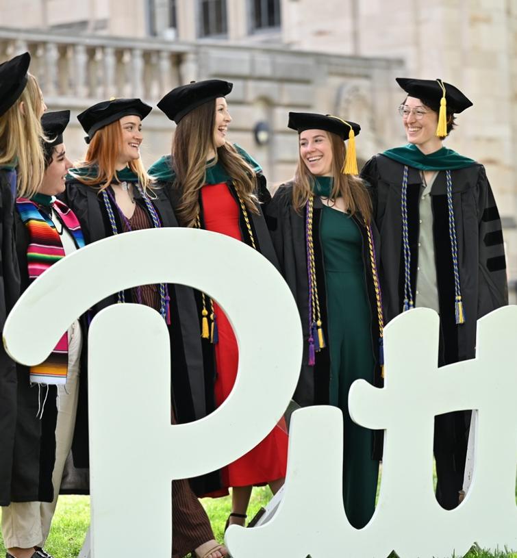 Students in caps and gowns stand behind a wood cutout that says, "Pitt"
